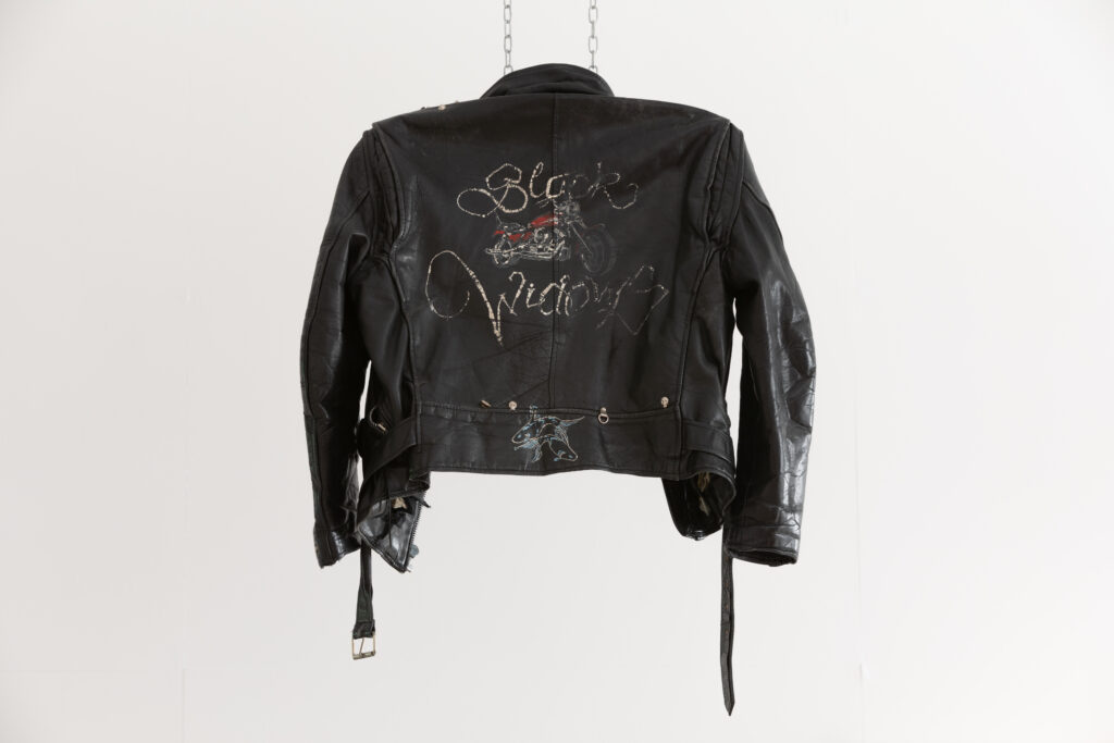 Leather jacket hung from ceiling with chains. Black widows logo with words and motorbike painted on the back, worn. Rebel Dykes - Fisch - Black Widows Leather Jacket