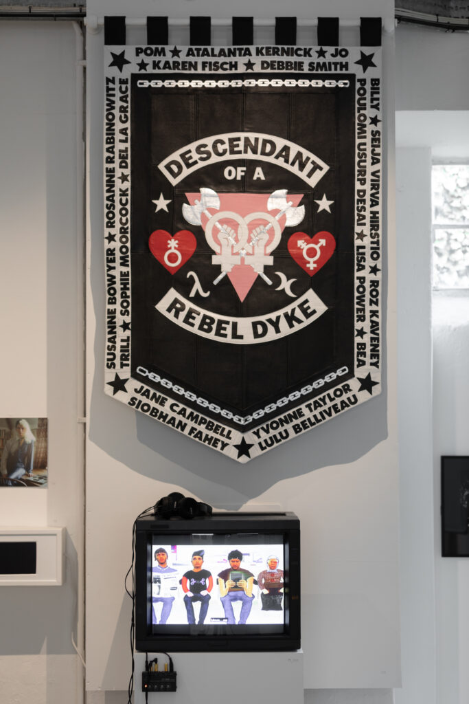 Leather banner reads 'Dependent of a Rebel Dyke' around a central motif of interlocking double female sign with fists of power holding labryes on top of upside down light pink triangle, framed by genderqueer and transgender symbols in hearts. The names of rebel dykes featured in the documentary film around the boarder with chains and stars. Rebel Dykes - Emily Witham - Descendant of a Rebel Dyke, 2021 - Screen below shows video work by Kate Jessop - Kai Fiain installation also visible to left